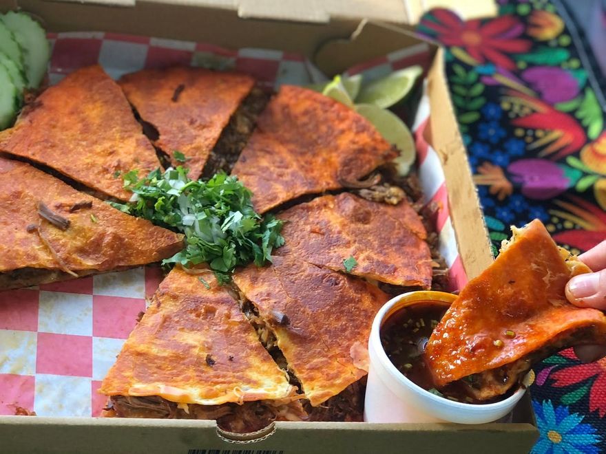 Pizza birria cut into slices in an open box, served with a cup of consommé for dipping.