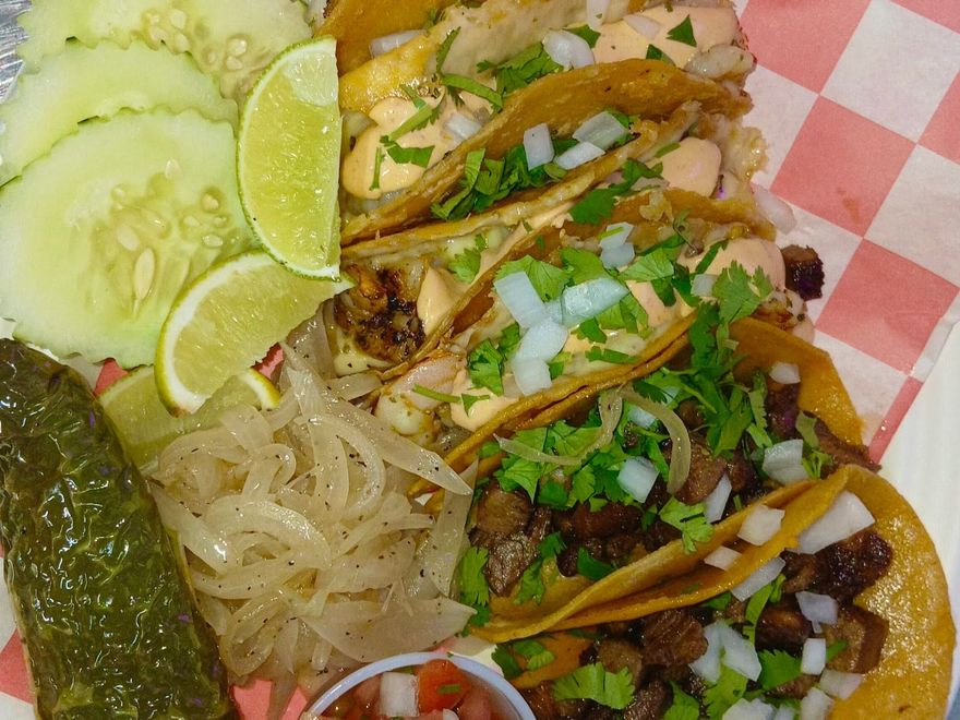 A lineup of corn-tortilla street tacos topped with onion and cilantro, with pico de gallo, lime, and grilled jalapeño on the side.